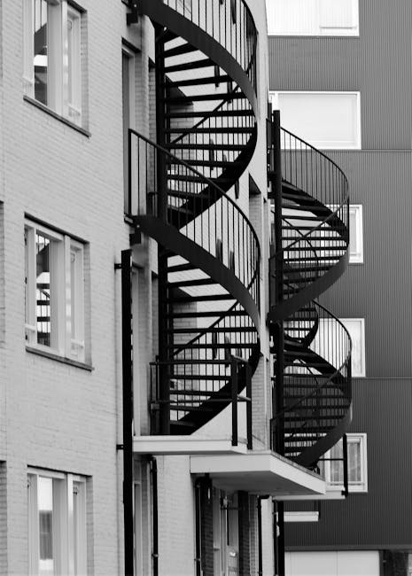 Interior view of a narrow stairwell in a Hackney flat showing black, textured carpeted steps with visible dust and dirt on their surfaces. A wooden handrail runs along the white tiled wall to the righ