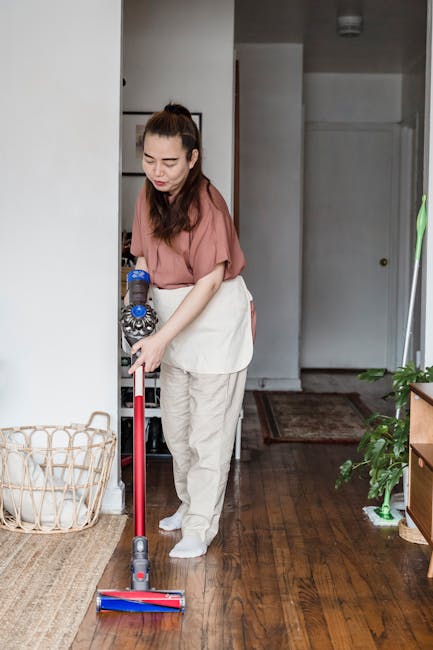 A woman cleaning a large glass window with a yellow cloth, using a spray bottle in her other hand. The window has multiple black metal frames and appears to be part of a modern interior, with white cu