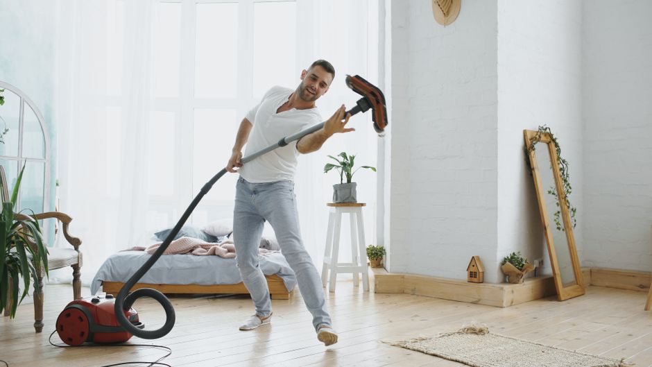 Close-up view of a vacuum cleaner with a transparent dust canister filled with debris, connected to a flexible, patterned hose attached to a floor brush. The vacuum is positioned on a pink carpeted fl